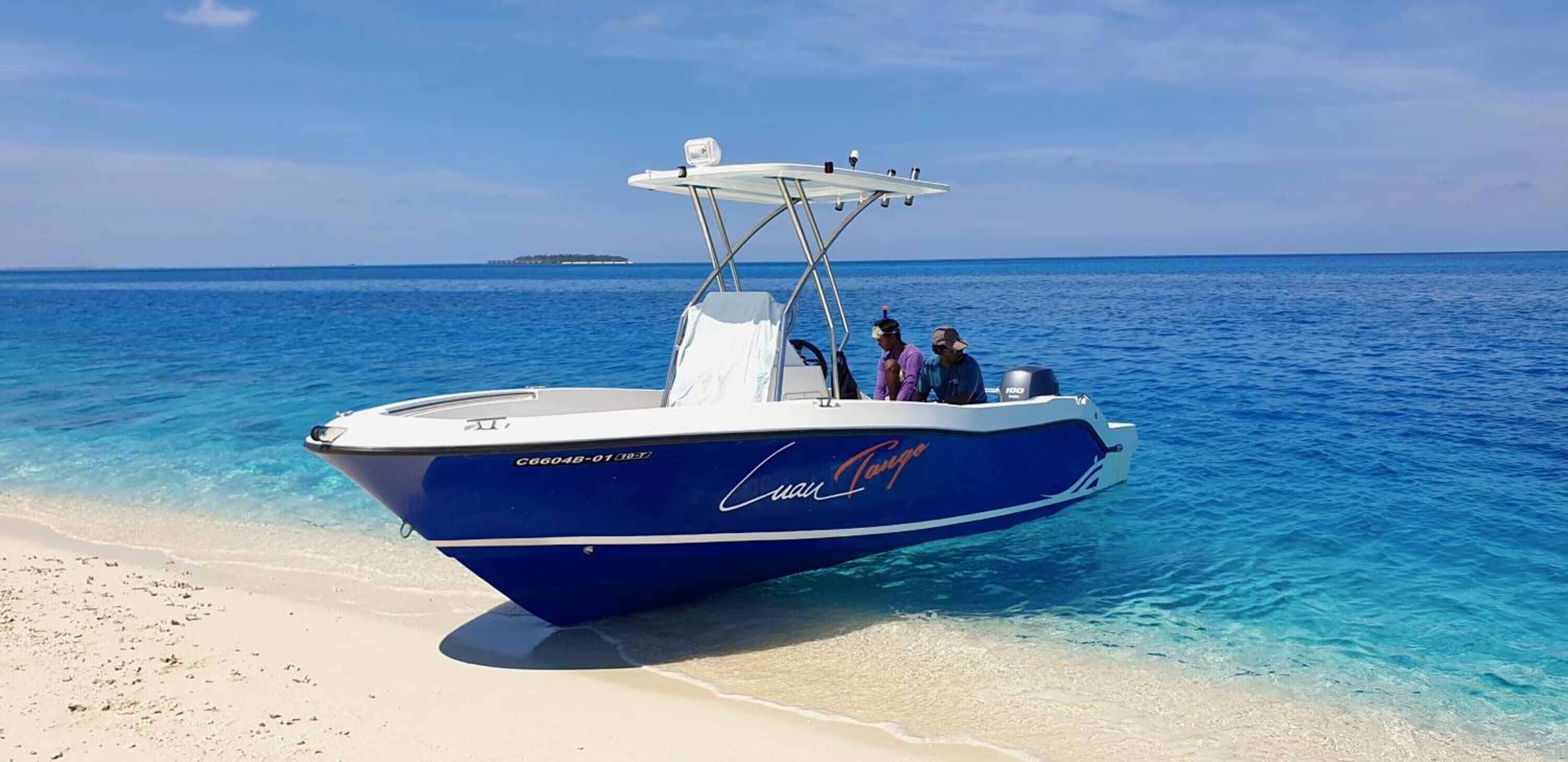 A boat named 'Cuan Tano' anchored near a beach with clear turquoise water and a distant island.