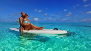 A couple sitting on a surfboard in clear turquoise water with a boat in the background. - Luau Beach Inn (Fulidhoo)