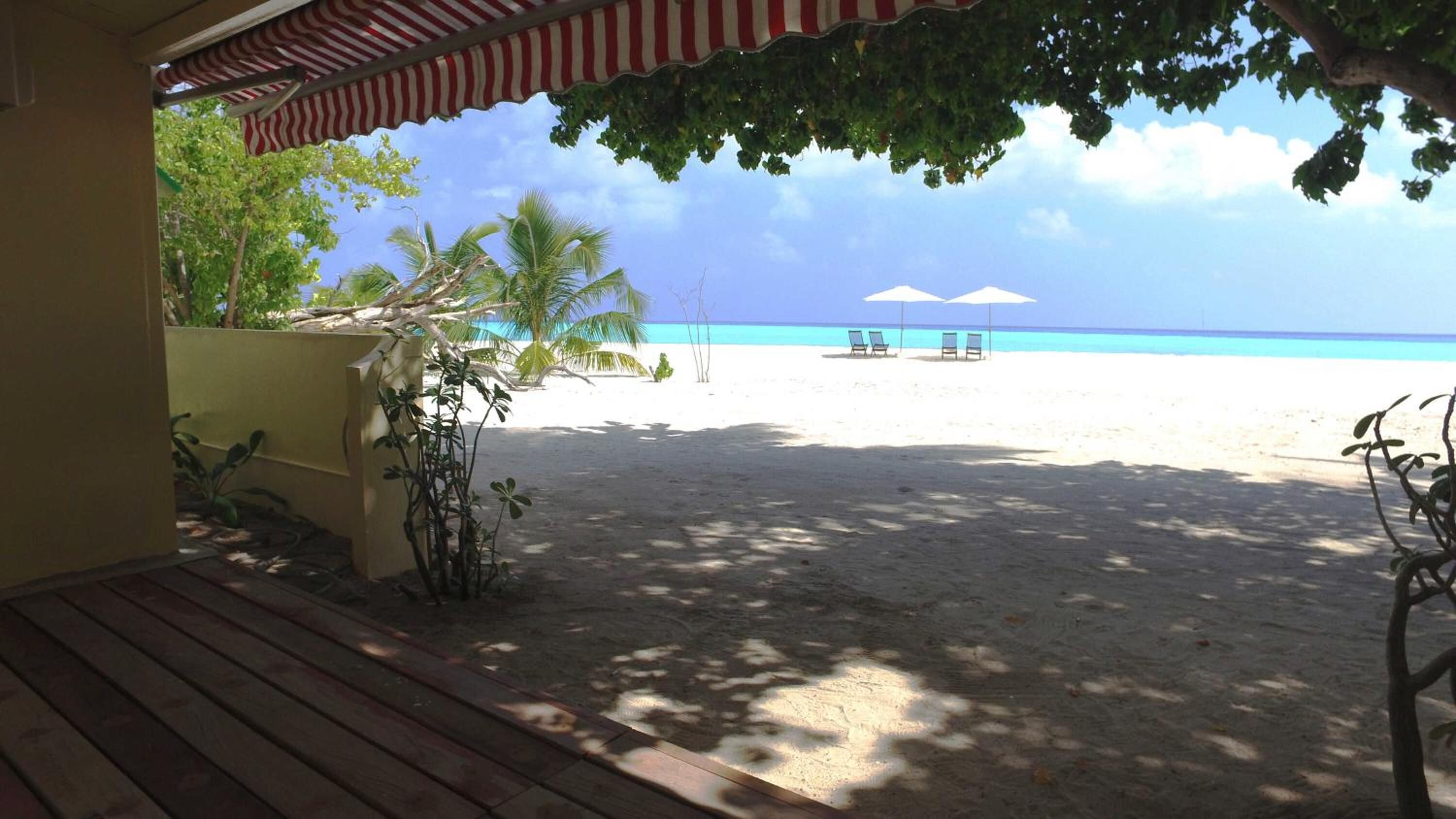 A beach view from a covered area with a wooden deck, two beach chairs, and umbrellas.