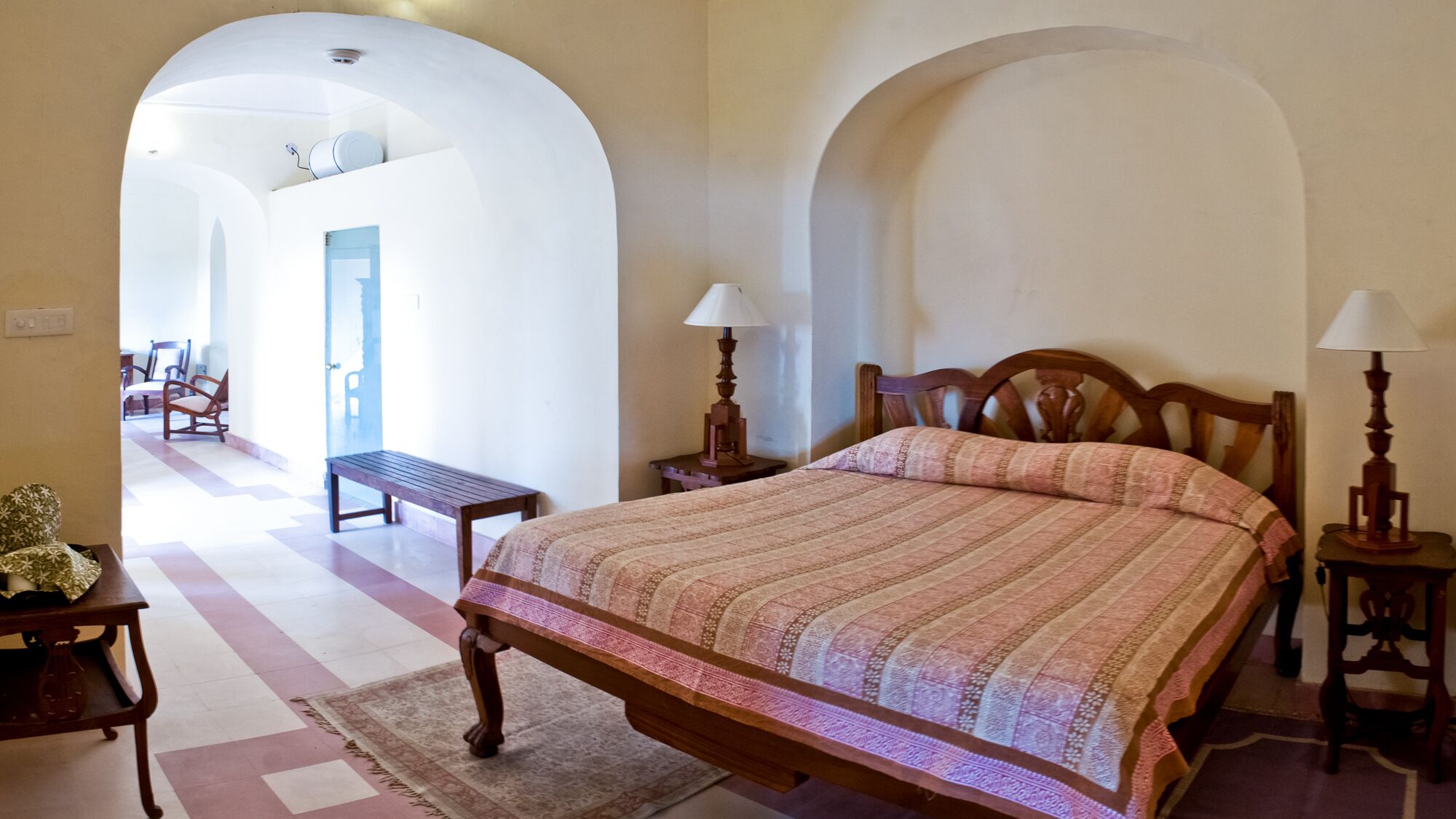 Un dormitorio con una cama grande, dos mesitas de noche con lámparas, un banco de madera y una alfombra estampada.