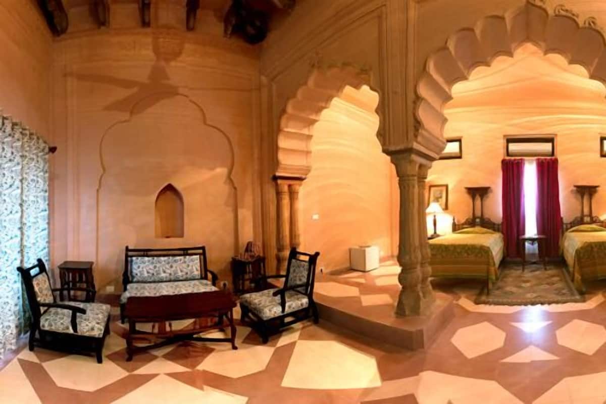 A room with a geometric patterned floor, a bed with a green bedspread, two chairs, a small table, and a large window with curtains.
