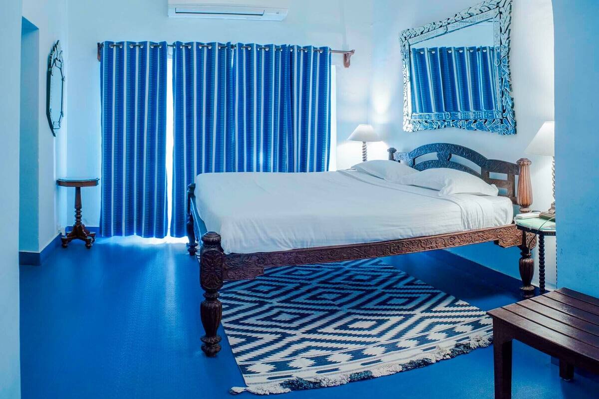 A bedroom with a large bed, a patterned rug, a wooden nightstand, and a mirror on the wall.