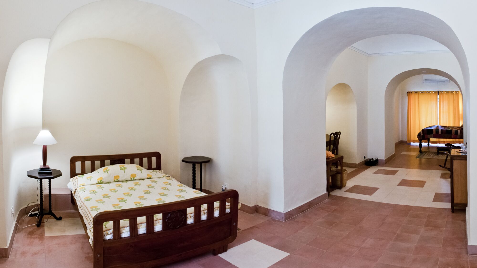 Quarto com cama de madeira, mesa de cabeceira com abajur, e vista para uma sala de jantar com mesa e cadeiras.