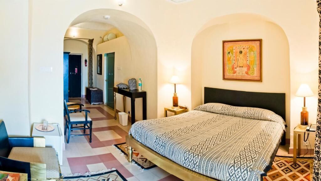 Ein Hotelzimmer mit einem Bett, zwei Nachttischen, einem Sessel, einem Schreibtisch, einer Lampe und einem Rundbogen.