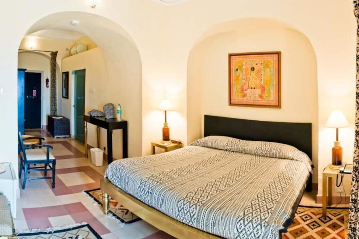 A hotel room with a bed, two bedside tables, a chair, a desk, a lamp, and an arched doorway.