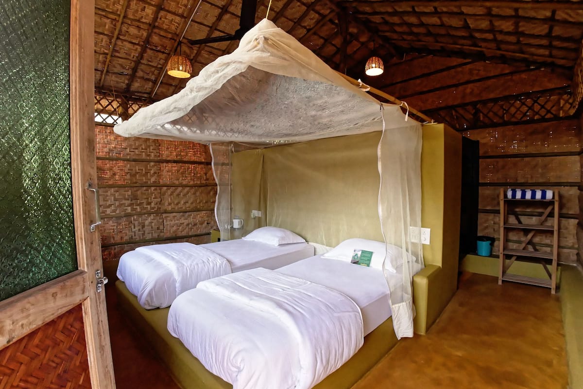 A room with two beds, a mosquito net, wooden walls, a woven ceiling, and a small shelf with a blue and white striped item.