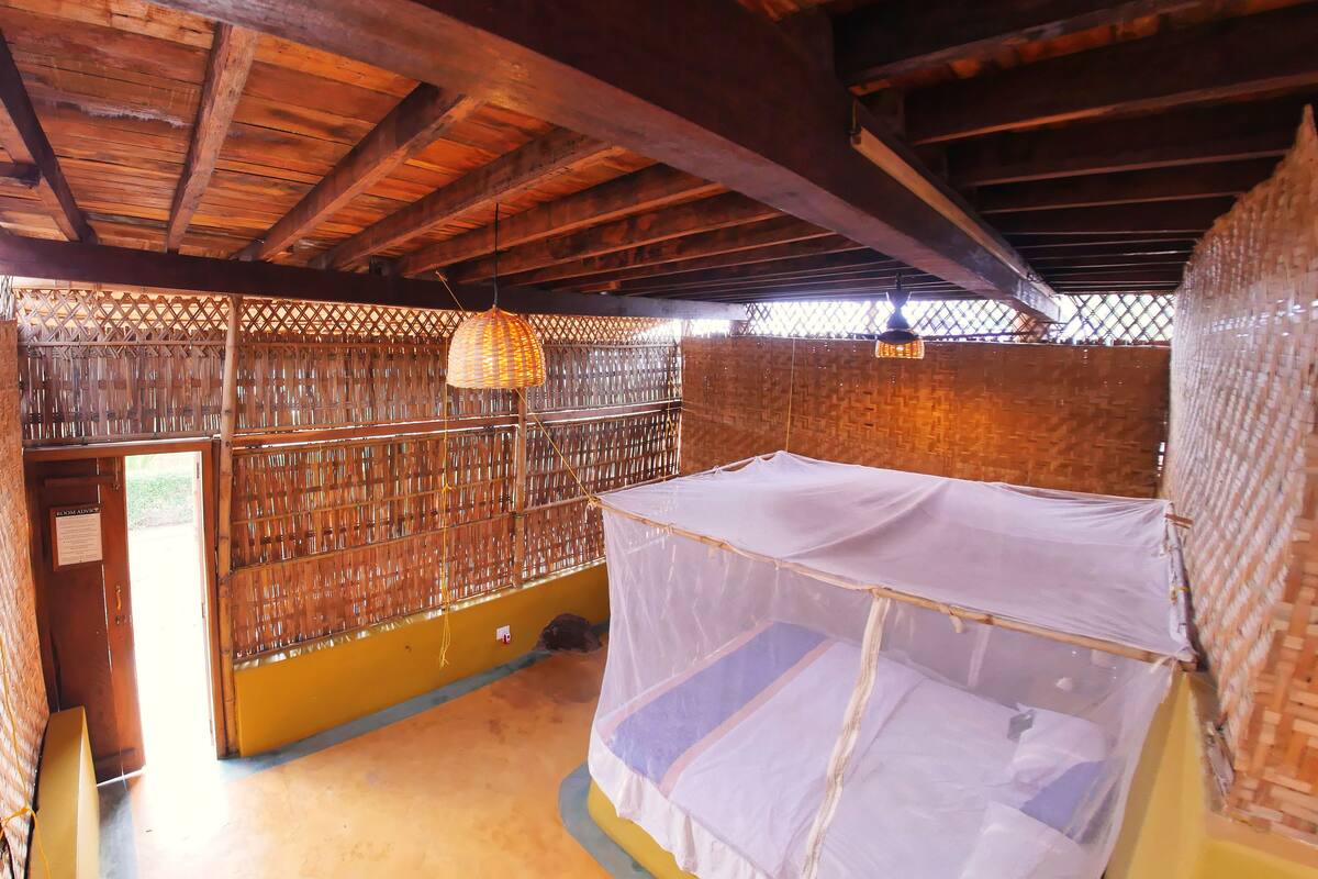 A room with a bed covered by a mosquito net, wooden beams on the ceiling, and a woven partition wall.