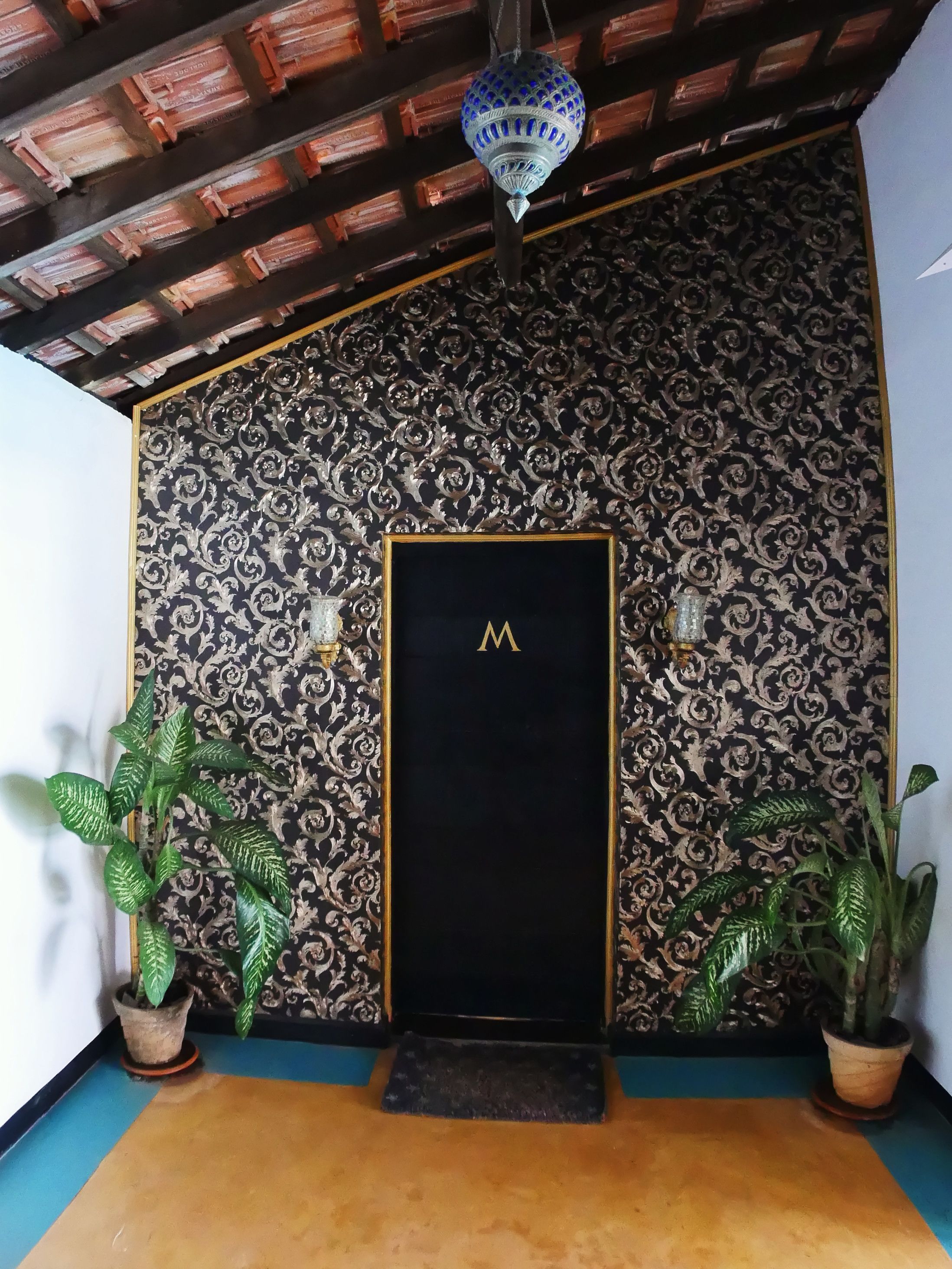 Una habitación con pared de diseño, una puerta negra con la letra 'M' dorada, dos plantas en macetas y una lámpara colgante.