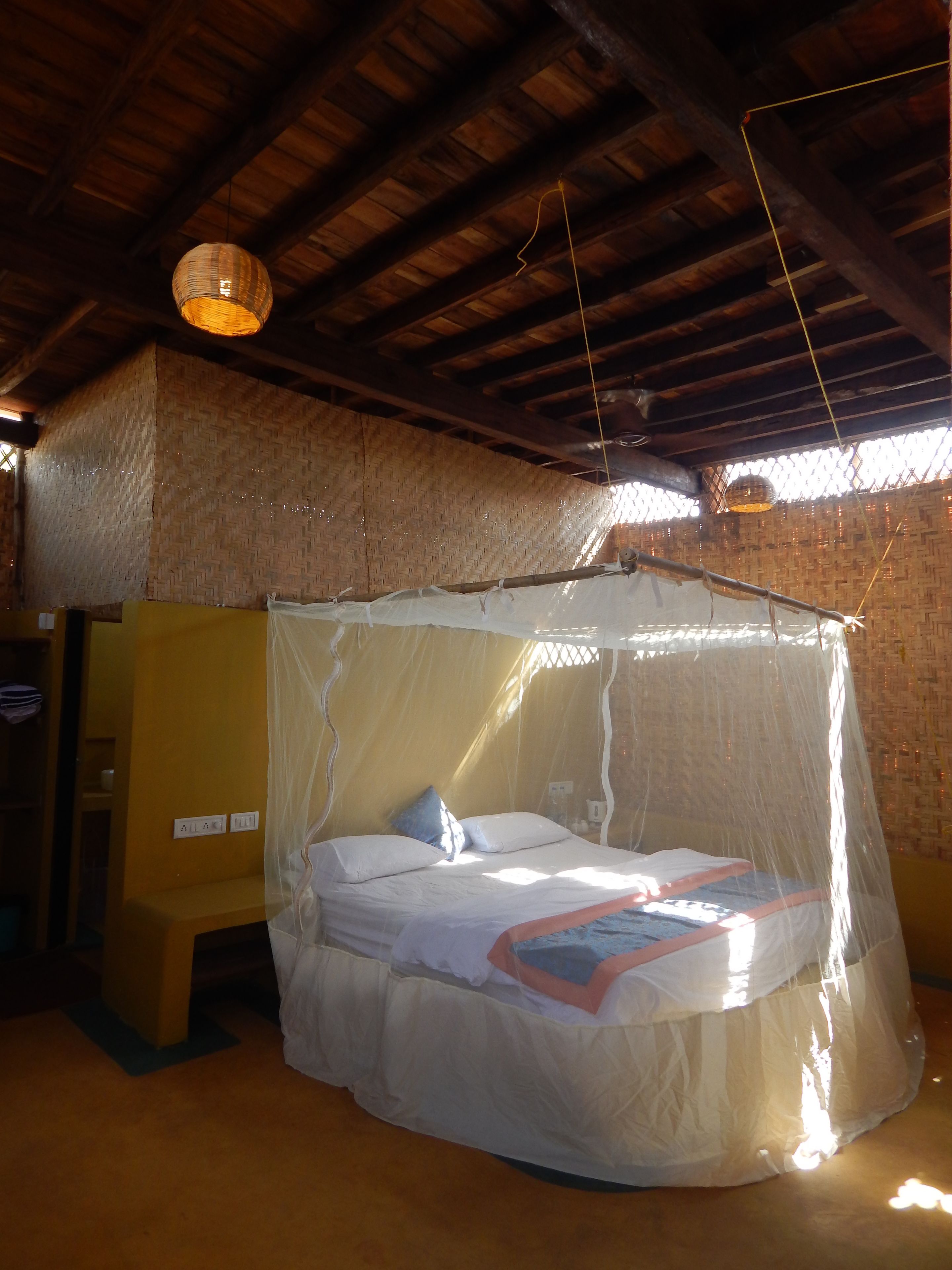 A room with a bed under a mosquito net, wooden ceiling beams, and a woven wall.
