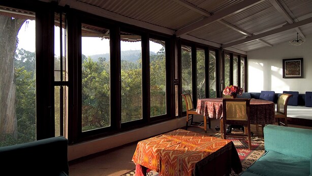 A room with large windows, a dining table with a patterned tablecloth, a sofa, and a chair.
