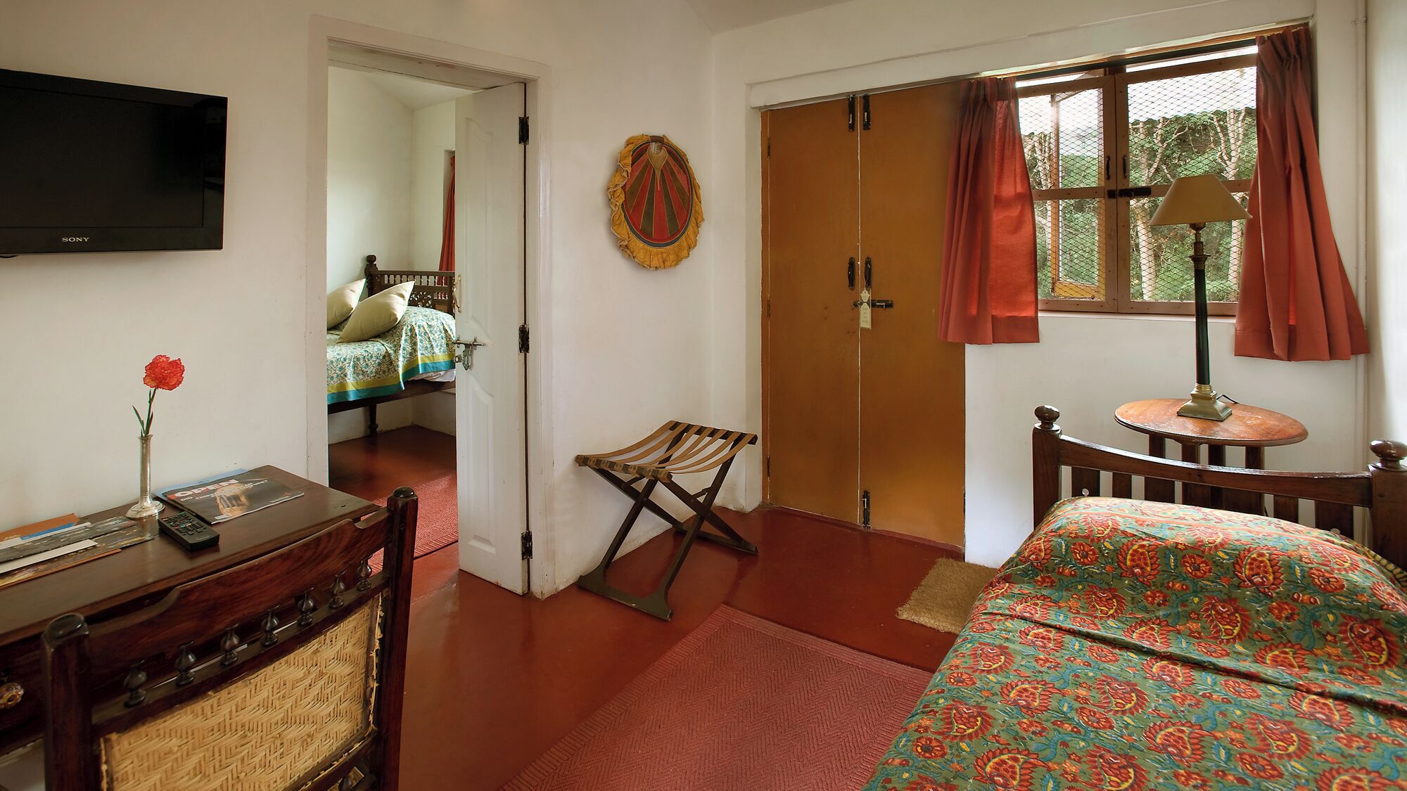 A room with a bed, a desk with a vase and remote, a chair, a door, and a window with curtains.