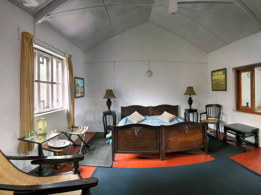 A bedroom with a wooden bed, two bedside tables with lamps, a chair, a small table with a kettle, and a window with curtains.