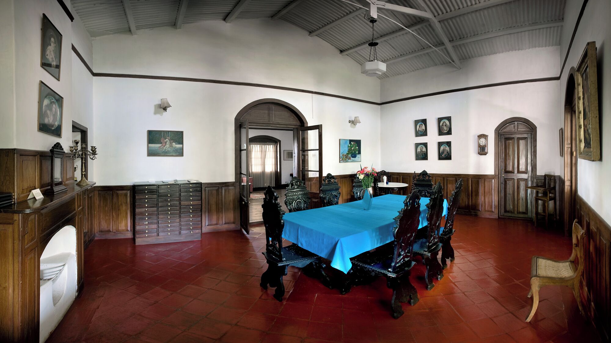 A dining room with a long table, wooden chairs, and a blue tablecloth. There are framed pictures on the walls and a clock on the wall.