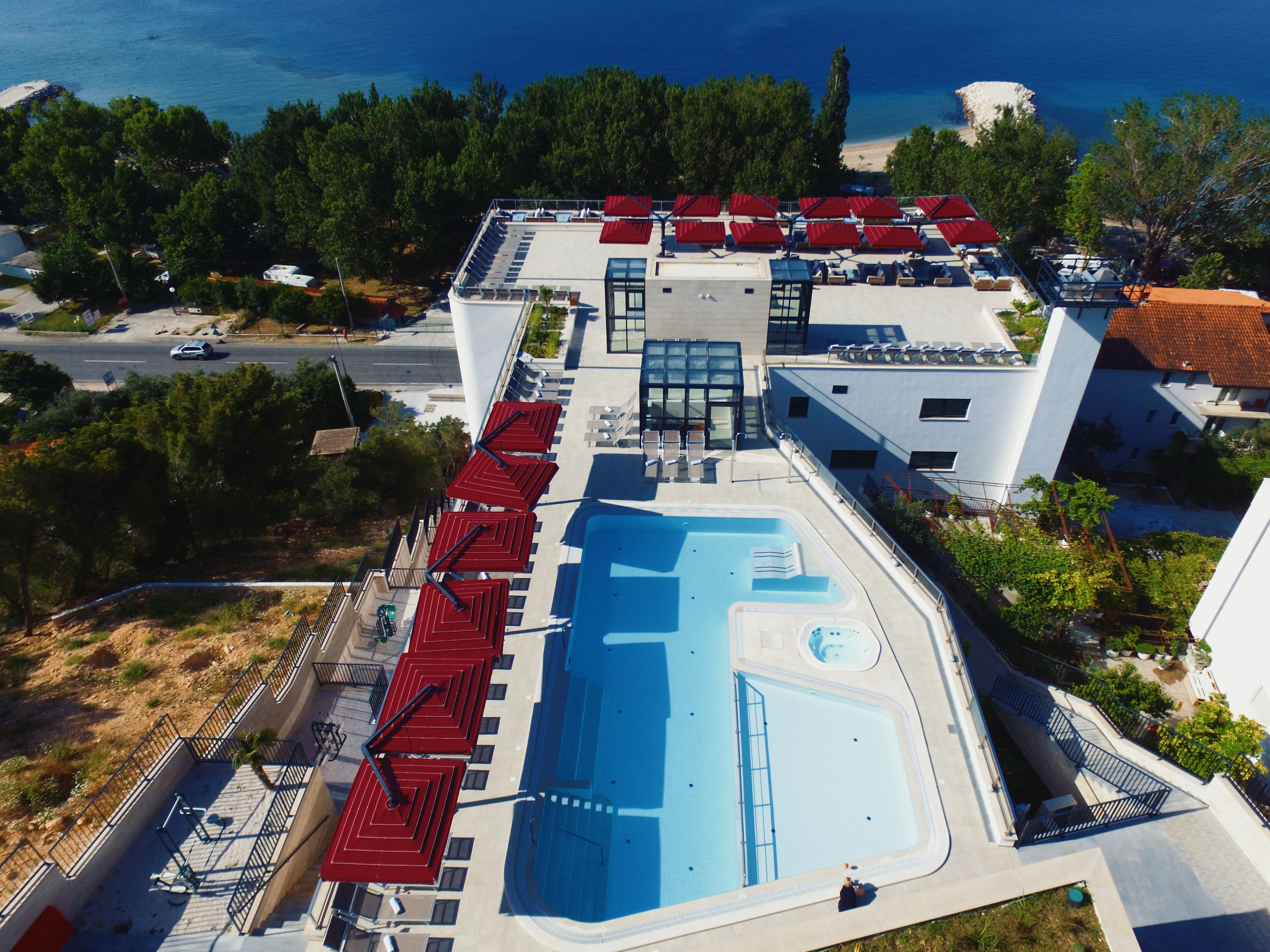 An aerial view of a hotel with a swimming pool, red umbrellas, and a view of the sea.