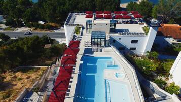 An aerial view of a hotel with a swimming pool, red umbrellas, and a view of the sea.