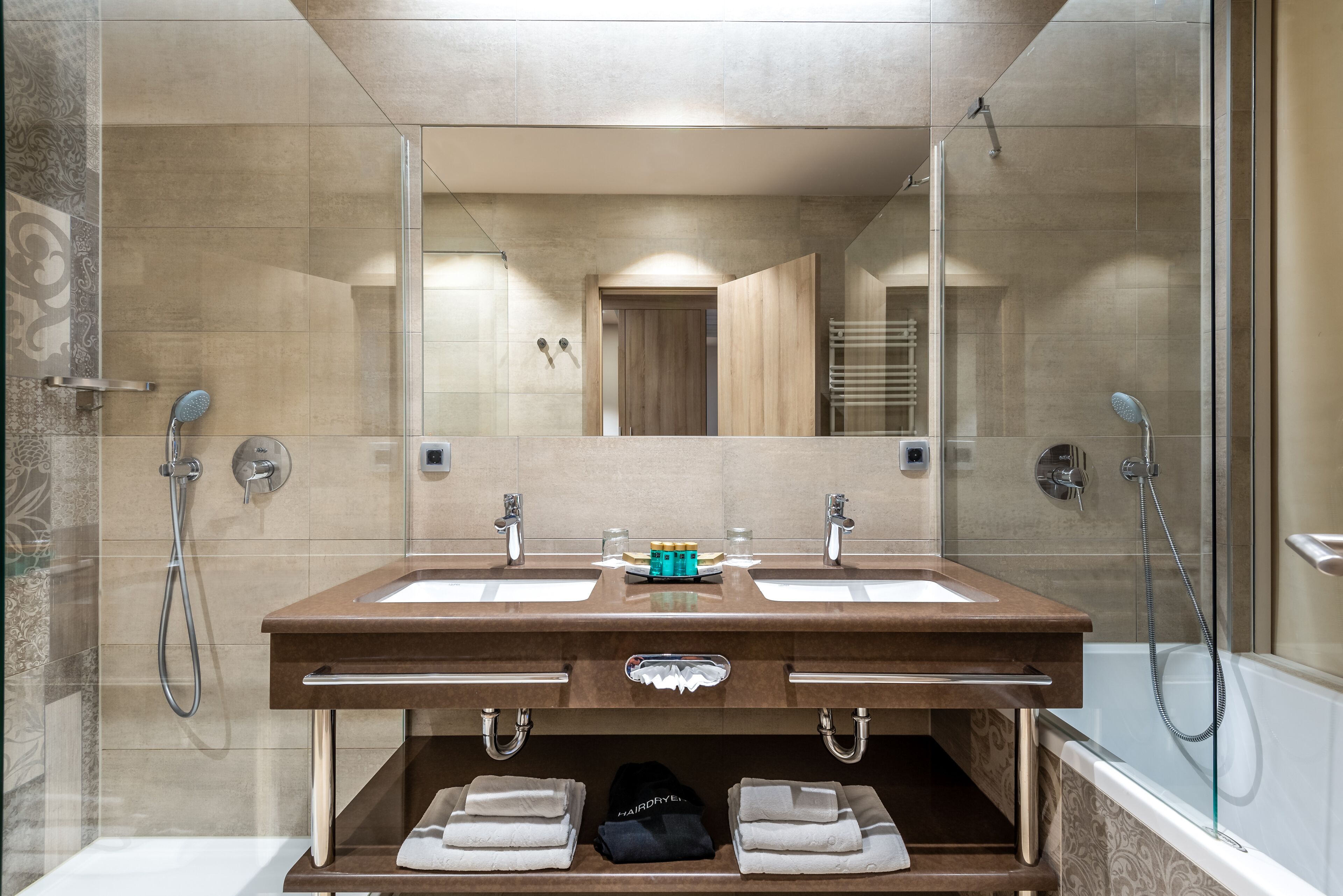 Een moderne badkamer met twee wastafels, een spiegel en een bad. Er liggen handdoeken en een zwarte tas op het aanrecht.