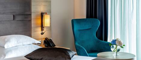 A hotel room with a bed, a nightstand with a lamp, a teal armchair, and a small table with a vase of flowers.