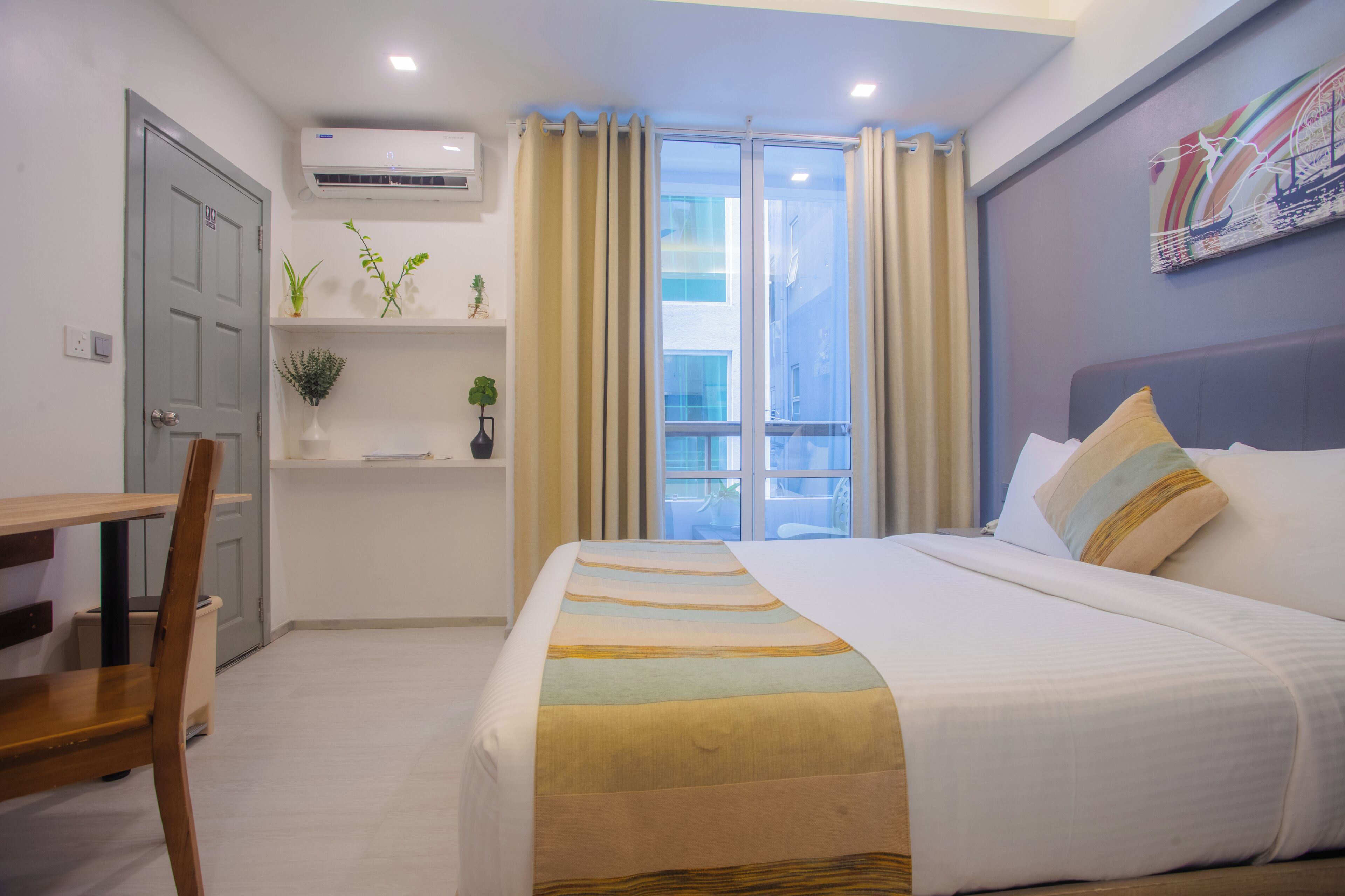 A hotel room with a bed, a desk, a chair, a window with curtains, a wall-mounted air conditioner, and a shelf with plants.
