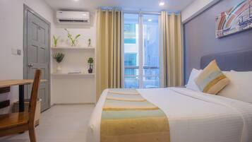 A hotel room with a bed, a desk, a chair, a window with curtains, a wall-mounted air conditioner, and a shelf with plants.
