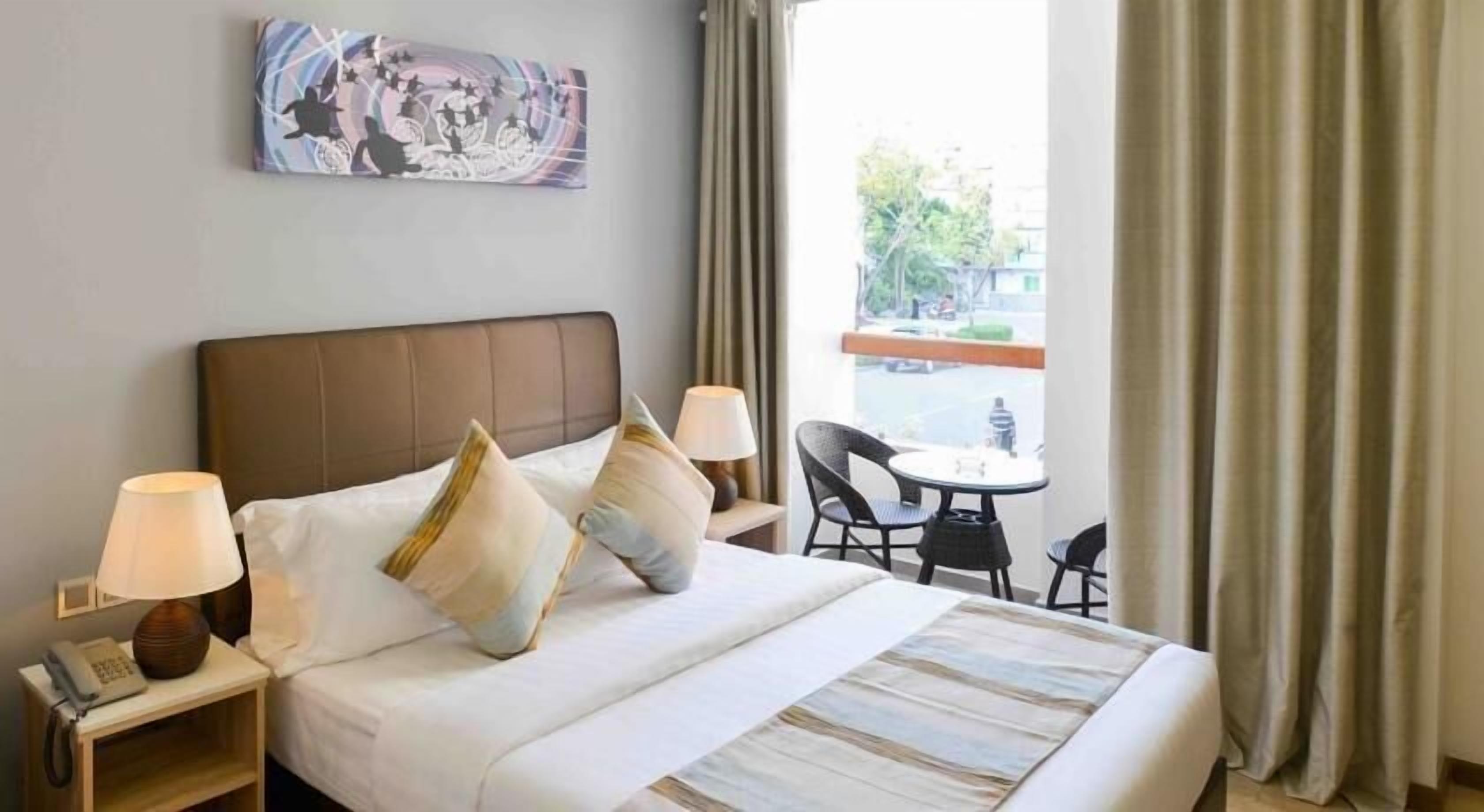 A hotel room with a large bed, two bedside tables with lamps, a small round table with two chairs, and a view of the outside through a window with curtains.
