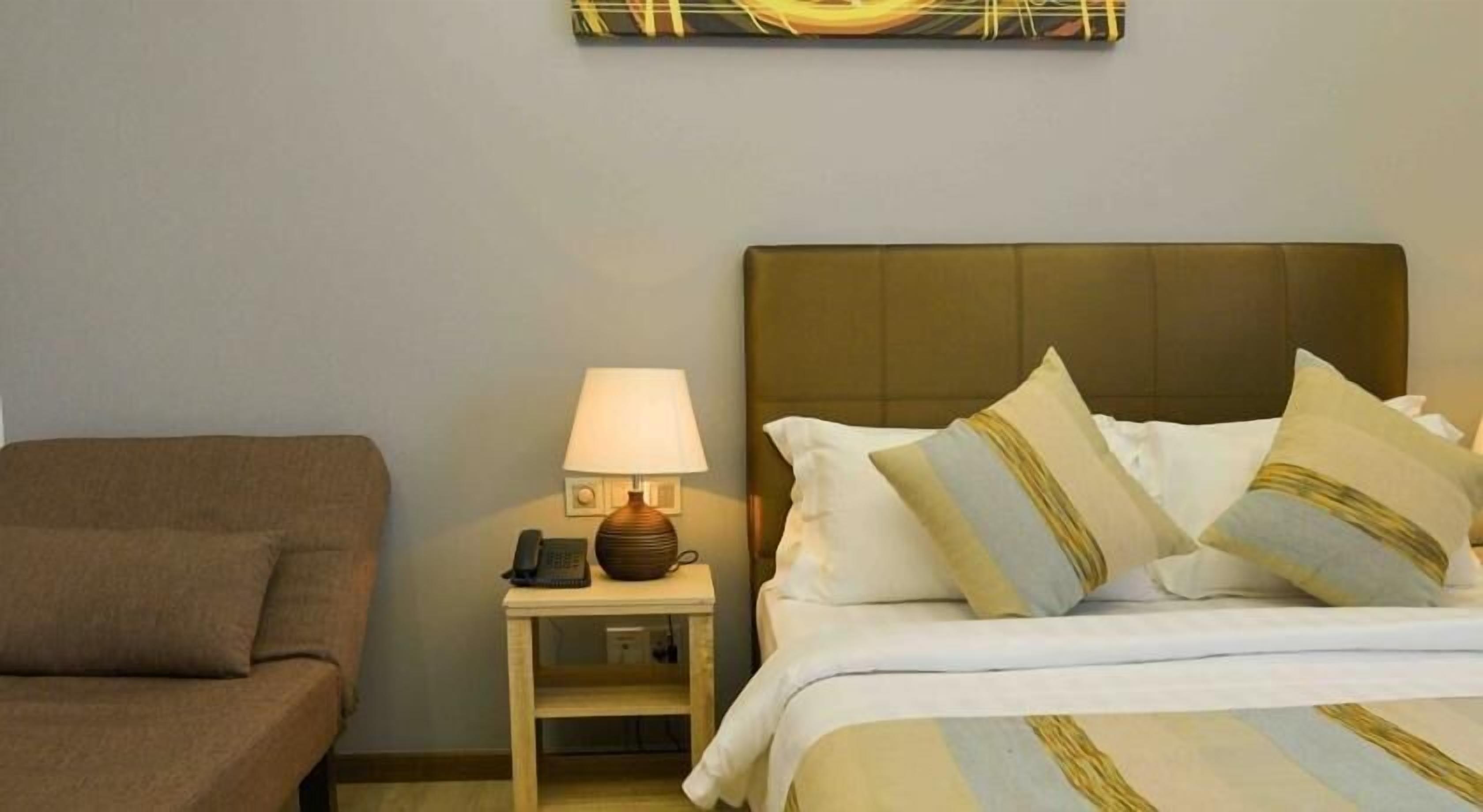 A hotel room with a bed, a nightstand with a lamp and phone, and a brown armchair.
