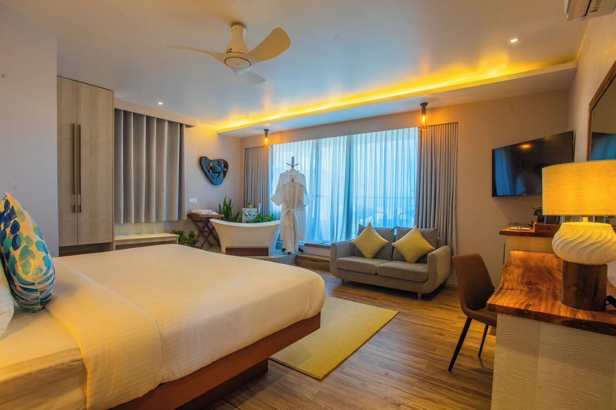 A modern hotel room with a large bed, a sofa, a desk with a lamp, a ceiling fan, and a TV.