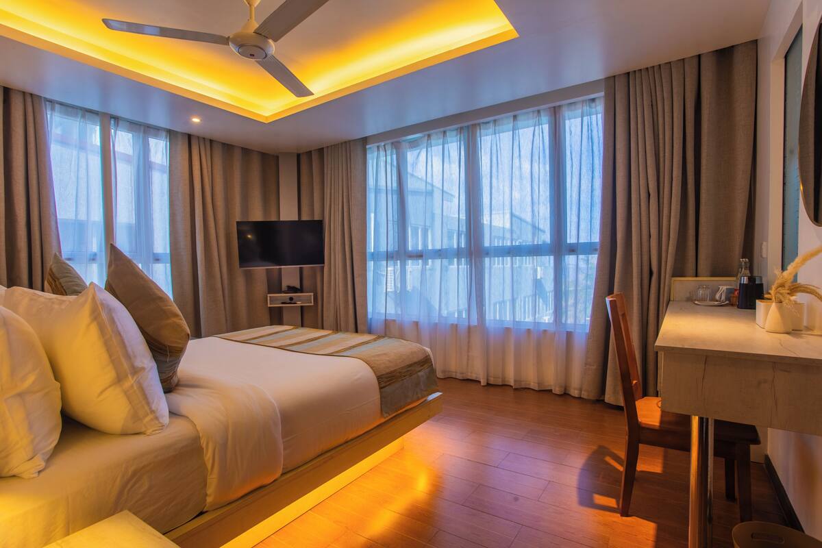 A hotel room with a bed, a desk, a chair, a television, and large windows with curtains.
