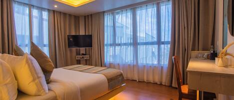 A hotel room with a bed, a desk, a chair, a television, and large windows with curtains.