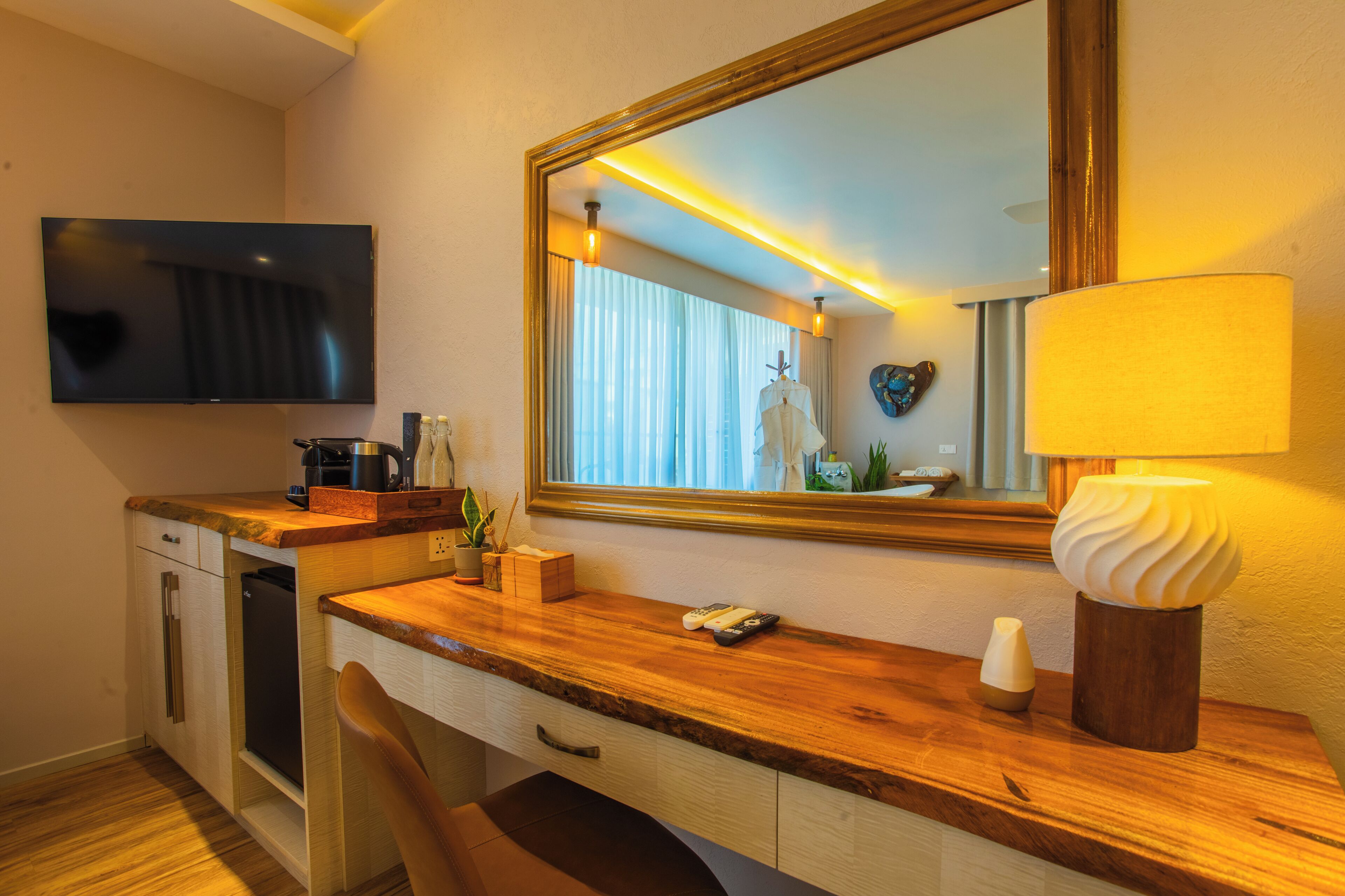 A hotel room with a wooden desk, a flat-screen TV, a mirror, a lamp, a plant, and a small refrigerator.