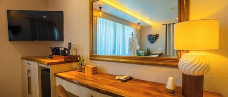 A hotel room with a wooden desk, a flat-screen TV, a mirror, a lamp, a plant, and a small refrigerator.