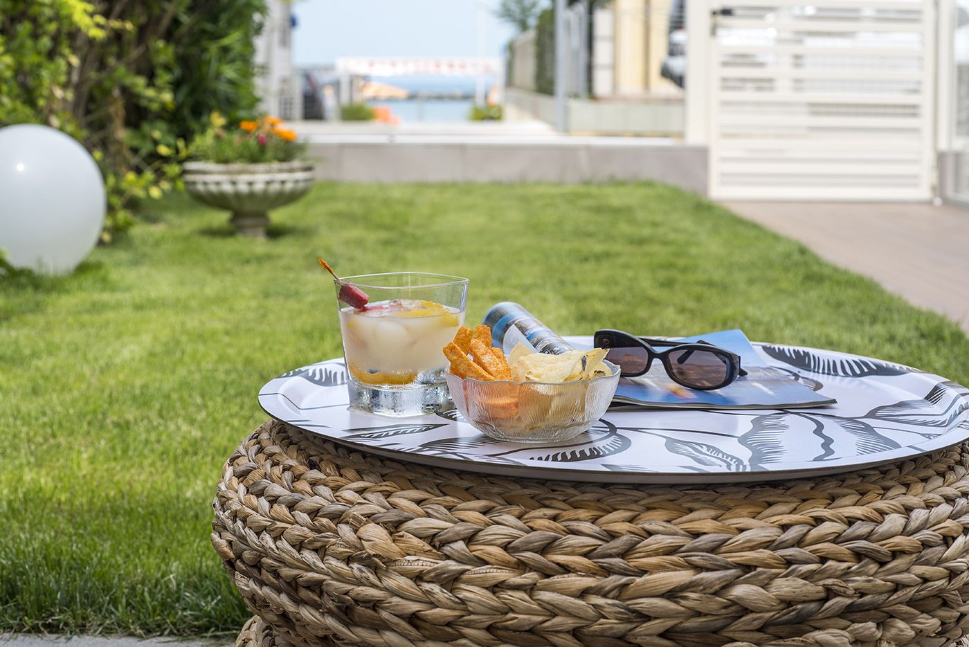 A wicker table with a glass of iced drink, a bowl of chips, and sunglasses on a grassy area with a view of a building in the background.