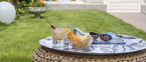 A wicker table with a glass of iced drink, a bowl of chips, and sunglasses on a grassy area with a view of a building in the background.