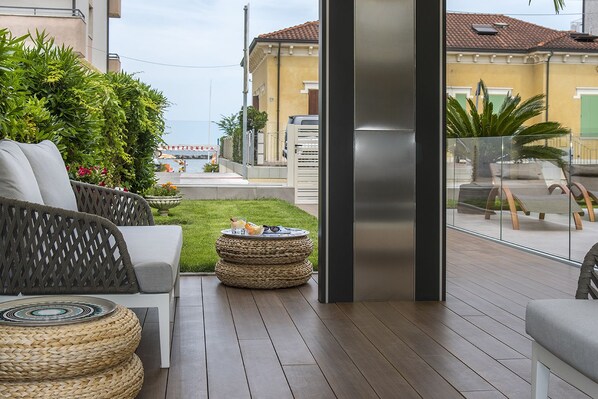 A modern outdoor patio with wicker furniture, a round woven table, and a view of a residential area with a yellow building. - Hotel Olimpia (Bellaria-Igea Marina)