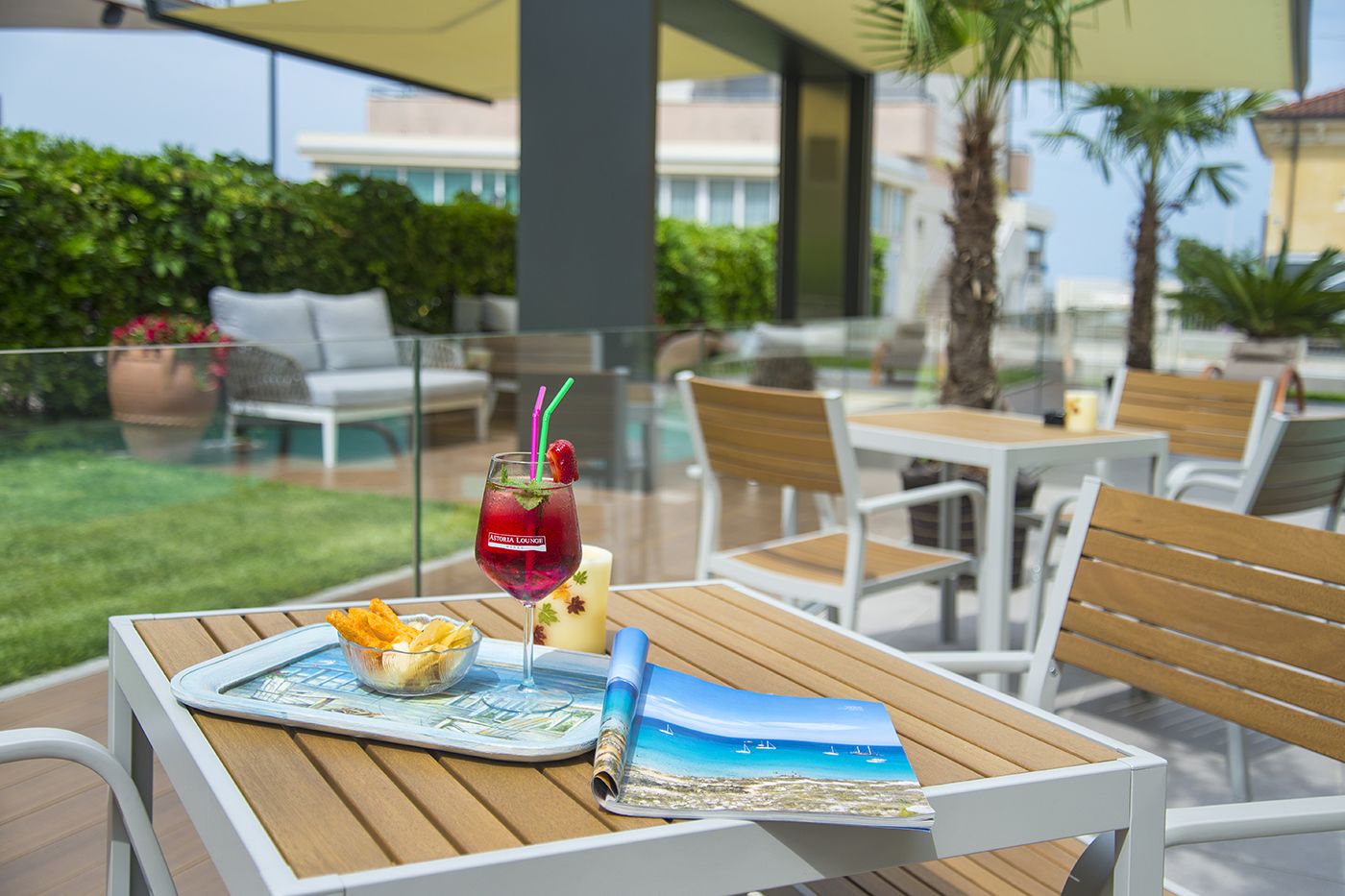 An outdoor seating area with wooden tables and chairs, a drink with a straw, a candle, and a magazine on a table.