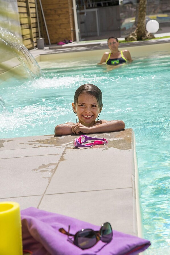 A pool area with a child resting on a pool edge, another person in the water, and a water slide in the background. - Hotel Olimpia (Bellaria-Igea Marina)