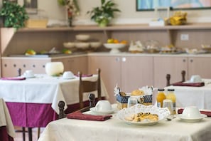 Une table dressée pour le petit-déjeuner, avec une assiette de viennoiseries, un panier de pain et deux bouteilles de jus d’orange.