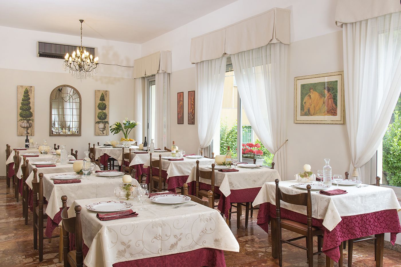 Zona pranzo con diversi tavoli apparecchiati, caratterizzata da un lampadario, ampie finestre con tende e opere d'arte alle pareti.