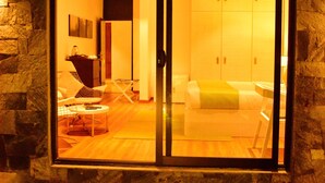 Un intérieur moderne avec un mur en pierre, une porte vitrée donnant sur une pièce comprenant un lit et une chaise, ainsi qu’une petite table.