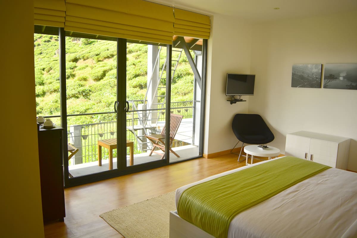 Un dormitorio con una cama grande, televisión, una mesita y una silla. Tiene un balcón con vistas a zonas verdes.