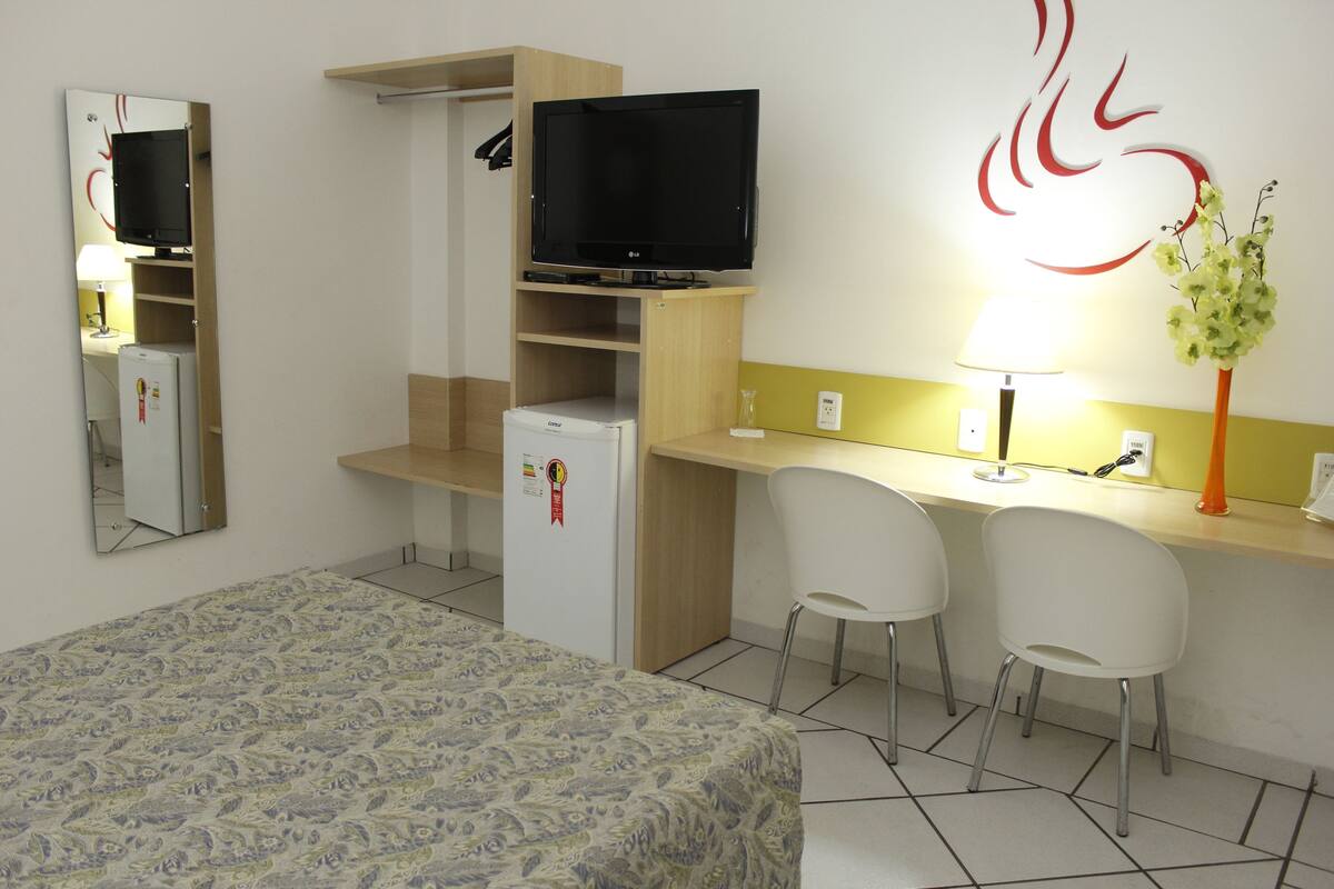 Una habitación de hotel con una cama, un escritorio con dos sillas, un televisor en un estante, un refrigerador y una decoración en la pared.