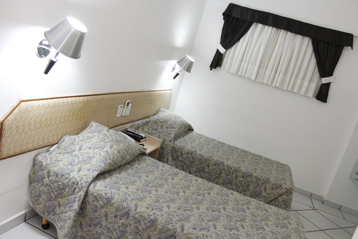 Habitación de hotel con dos camas, cabeceros, lámparas de noche, una ventana con cortinas y un teléfono en la pared.