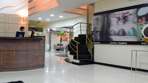 A hotel reception area with a wooden reception desk, a receptionist, a seating area with a couch and a vase of flowers, and a staircase leading to another floor. - Barao Center Hotel (Santarém)