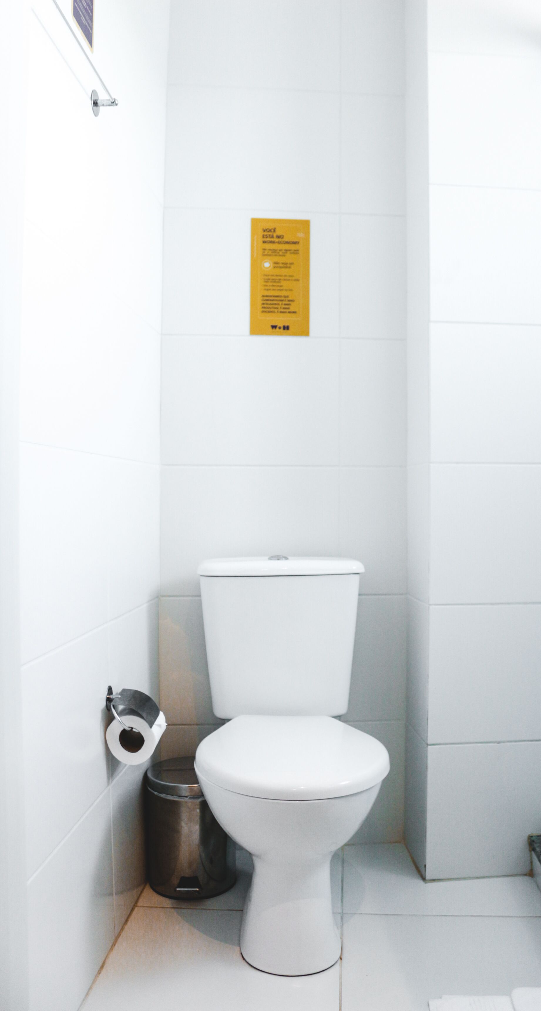 Inodoro blanco con tanque, un dispensador de papel higiénico y un cubo de basura en una esquina de un baño con azulejos blancos.