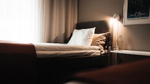 A neatly made bed with white and beige bedding, a bedside table with a lamp, and a framed picture on the wall. - Ädelfors Hotell och Konferens (Holsbybrunn)