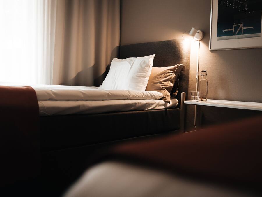 A neatly made bed with white and beige bedding, a bedside table with a lamp, and a framed picture on the wall.