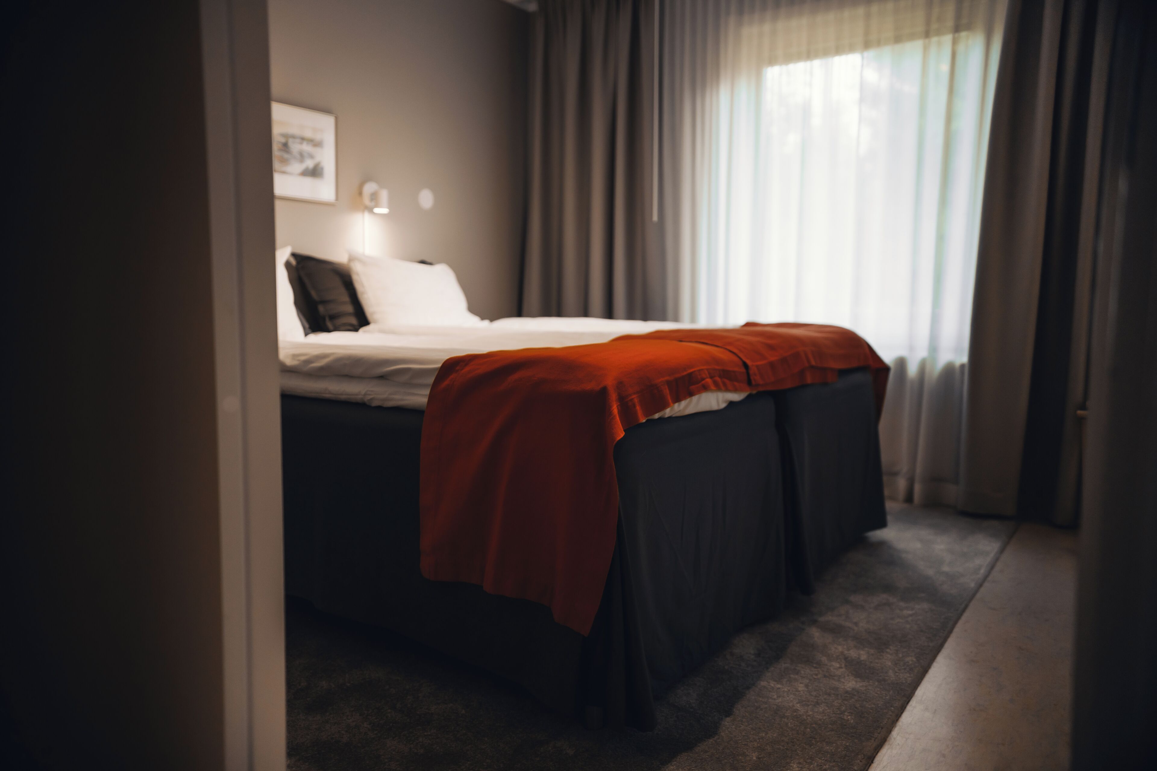 A neatly made bed with a red blanket, a bedside lamp, and a framed picture on the wall.