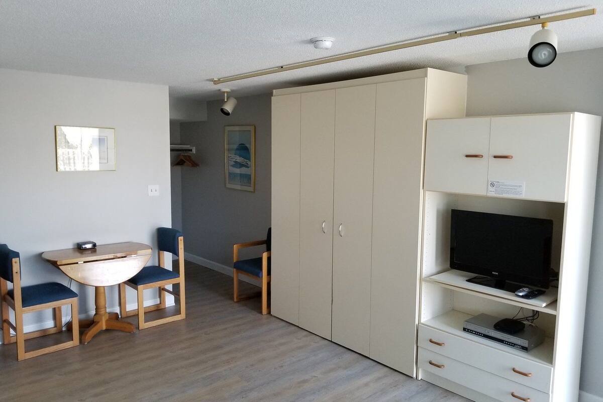 A room with a wooden dining table and chairs, a white wardrobe, and a TV on a shelf.