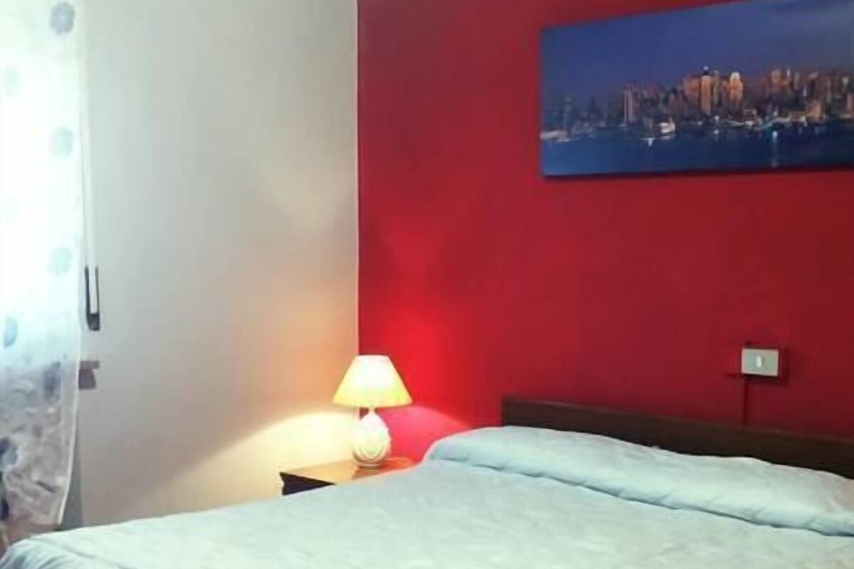 A room with a bed, a chair, a lamp, and a cityscape painting on the wall.