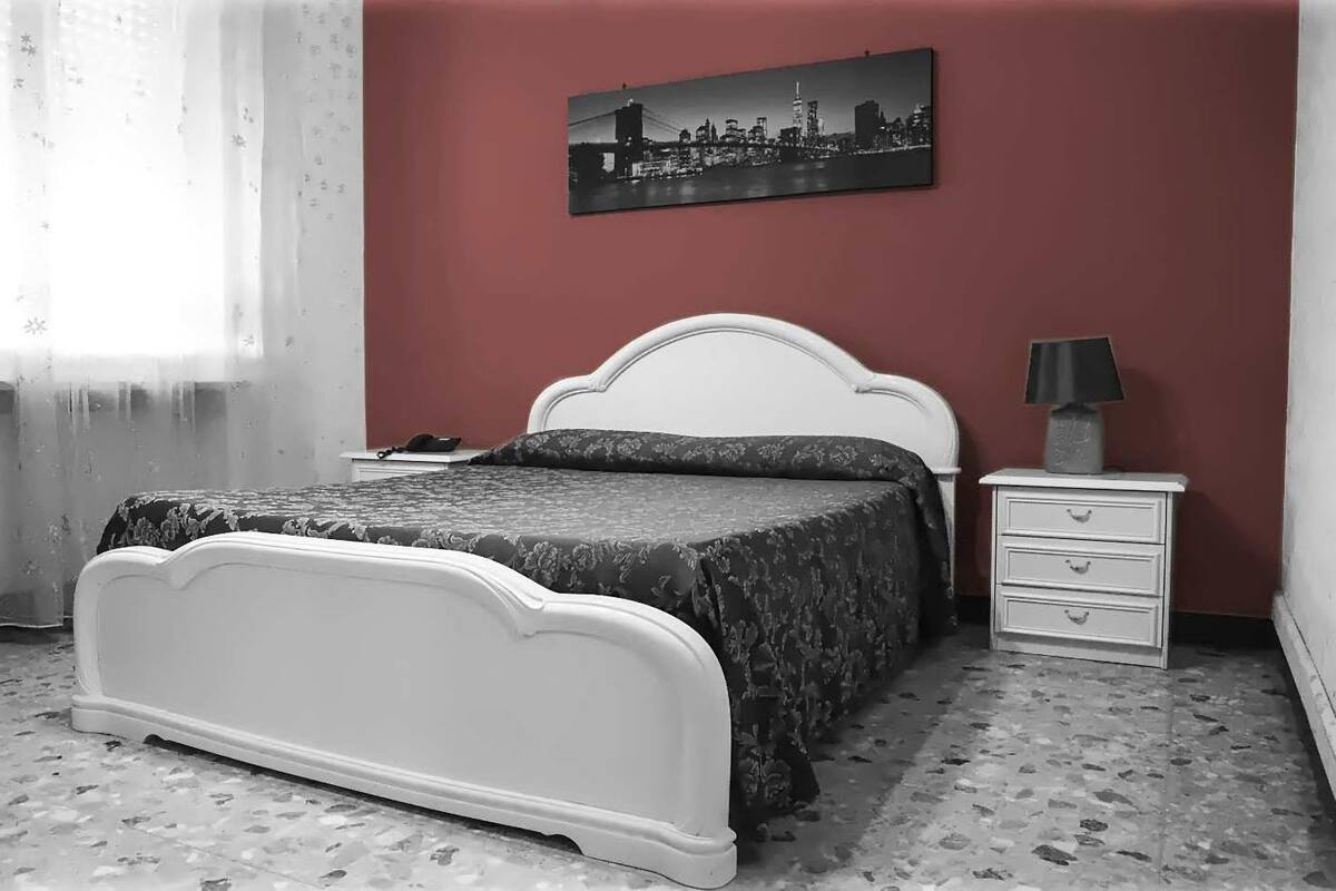 A bedroom with a white bed, a nightstand with a lamp, and a cityscape painting on the wall.