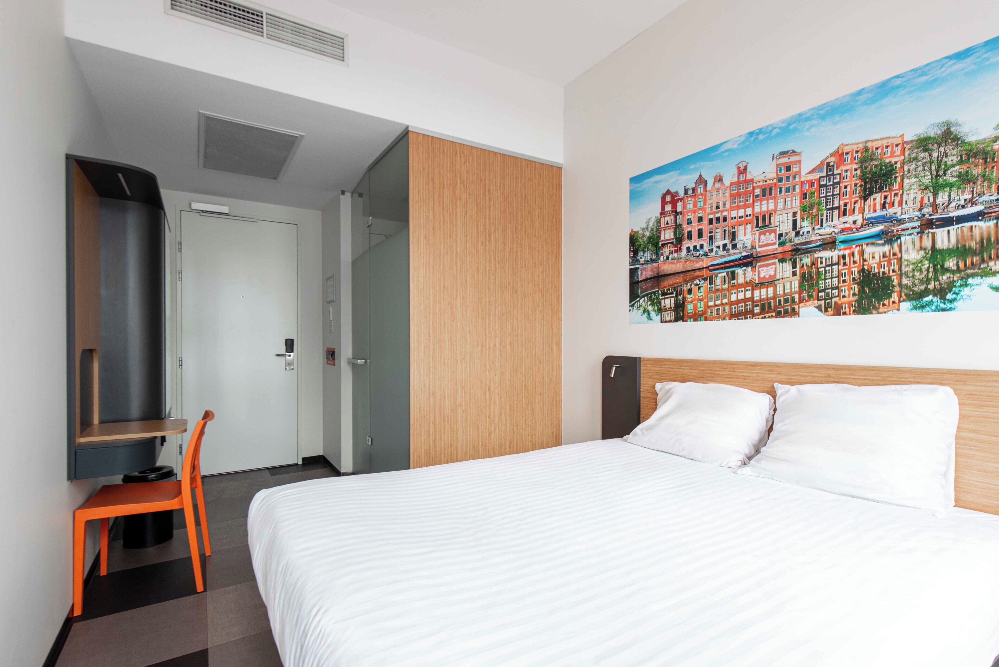 Habitación de hotel con cama, escritorio, silla, cuadro de un paisaje urbano y pared de paneles de madera.